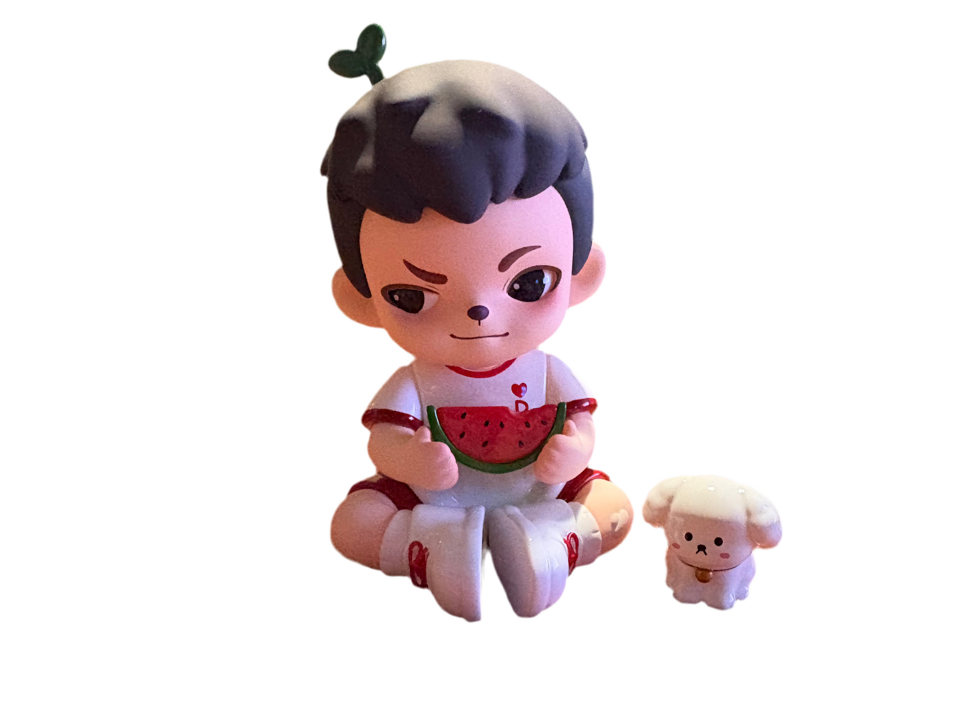 Toy figure of a boy with black hair, sitting and holding a watermelon slice, with a small dog toy next to it on a white background.